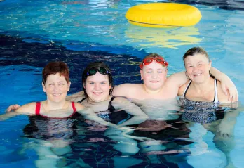 A family of four (two women, a girl and boy) smile and hug in a YMCA pool.