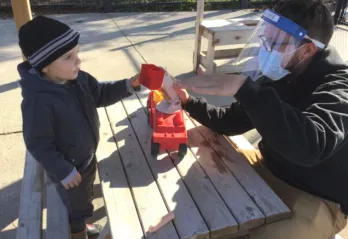 A young boy and Early Childhood Educator in PPE play at an outdoor picnic table with a red truck.