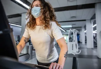 A woman with curly hair and tattoos on her arm, wearing a mask and white t-shirt, works out on a treadmill at the Y.