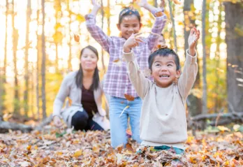 A young boy in a cream coloured sweater sits on the ground and smiles as he throws autumn leaves into the air. His sister and mom laugh and watch him from behind.