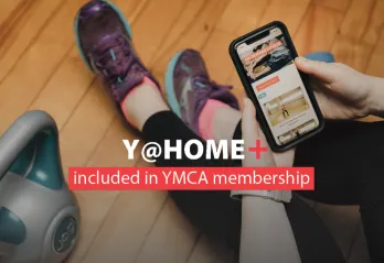 Y@HOME+ included in YMCA membership. Image: a woman in running shoes watches a workout on her mobile device next to a kettlebell.