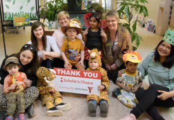 YMCA Staff pose with children from a Y Child Care Centre in costumes, holding a sign thanking Scholar's Choice for their sponsorship of Move-A-Thon.