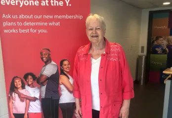 Barb smiles in front of a YMCA pop-up sign wearing a red shirt and black pants.