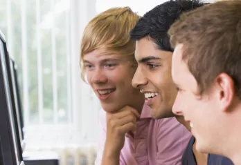 Three adolescents laugh as they play a game online in front of computer monitors.