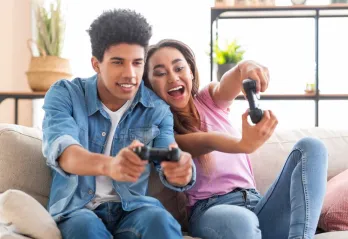 Two teens excitedly play video games on a couch.