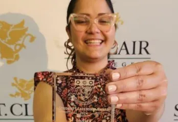 A smiling woman with glasses and dark hair pulled back holds up a clear glass award in front of her. The award reads "Student Leadership Award 2024" with the name "Indira" engraved below. She is wearing a floral dress, and the backdrop behind her features the logo and name of St. Clair College with gold griffin designs.