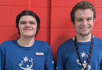 Two young men with light skin and short brown hair stand side by side in front of a red brick wall, smiling at the camera. They are both wearing matching blue t-shirts with a white logo featuring a stylized person, stars, and swirls. The man on the left has a name tag that reads 'Noah' and 'Project Search Team.' The man on the right wears a black lanyard with red 'Y' logos.
