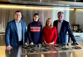 A group of four professionally dressed people stands behind a stainless steel kitchen counter with electric stovetops. They are smiling at the camera in a modern, well-lit kitchen with reflective metal surfaces, a large ventilation hood, and various kitchen appliances in the background.