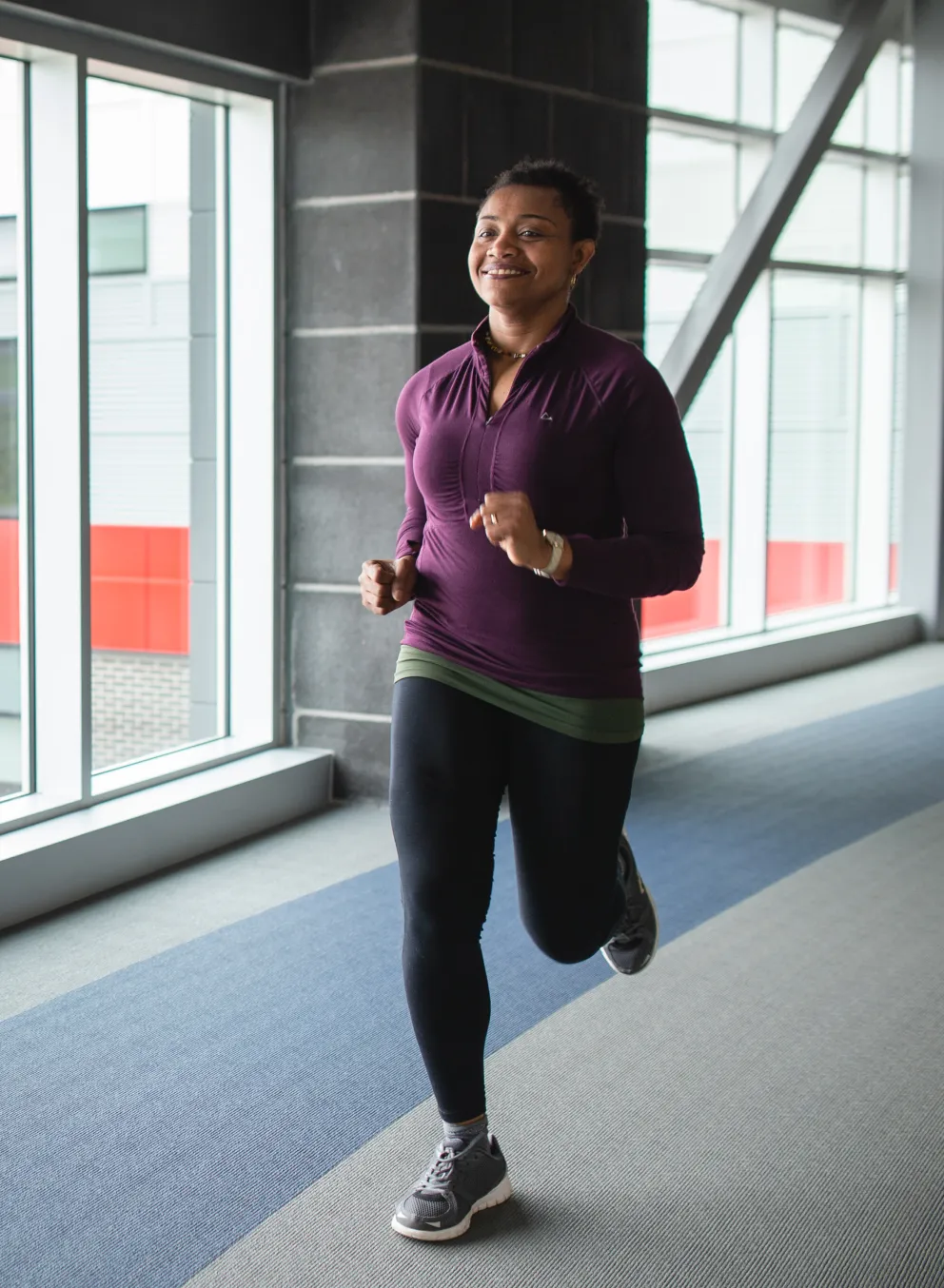 A woman in a purple shirt and black leggings jogs on an indoor walking track.