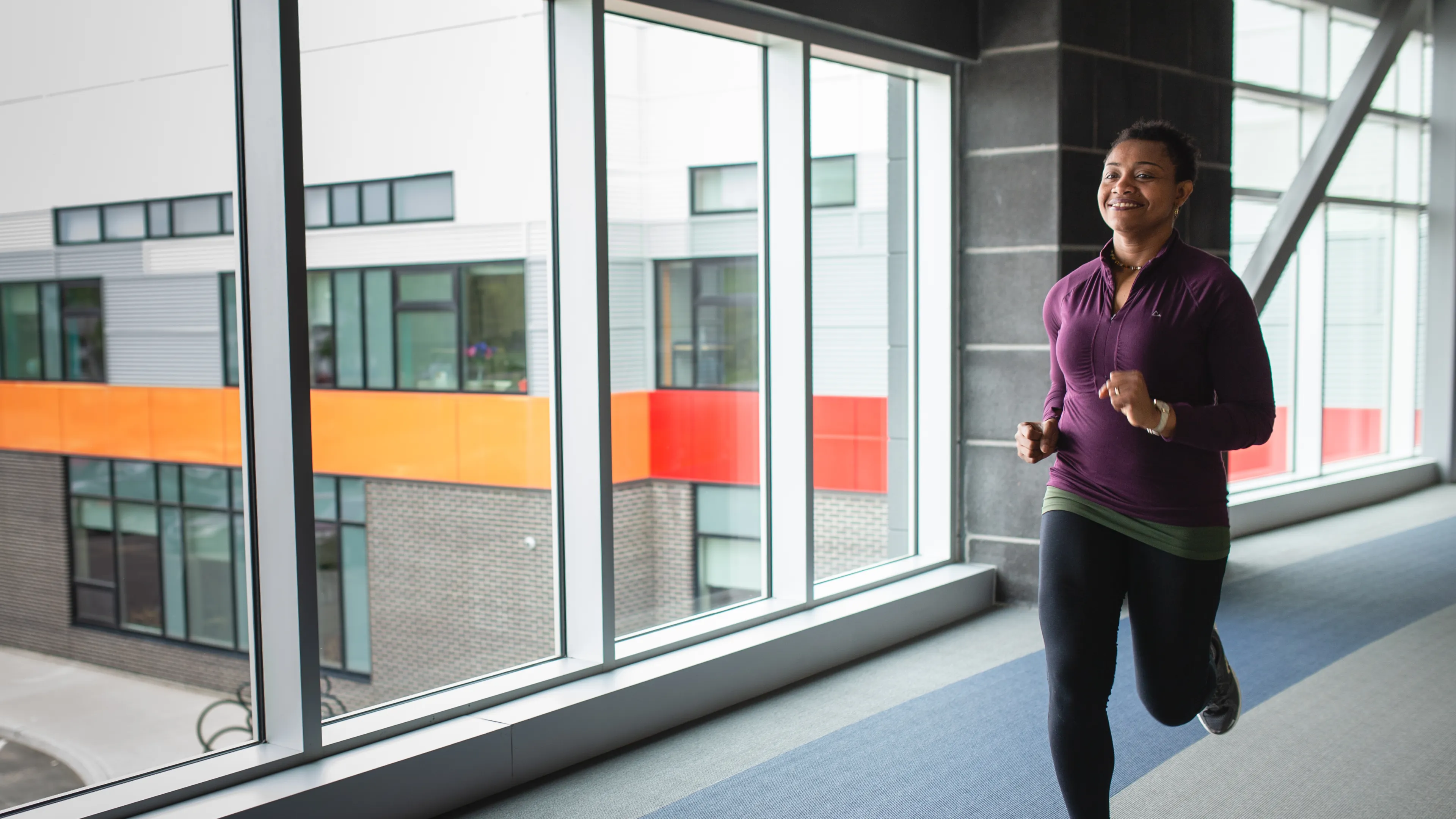 A woman in a purple shirt and black leggings jogs on an indoor walking track.
