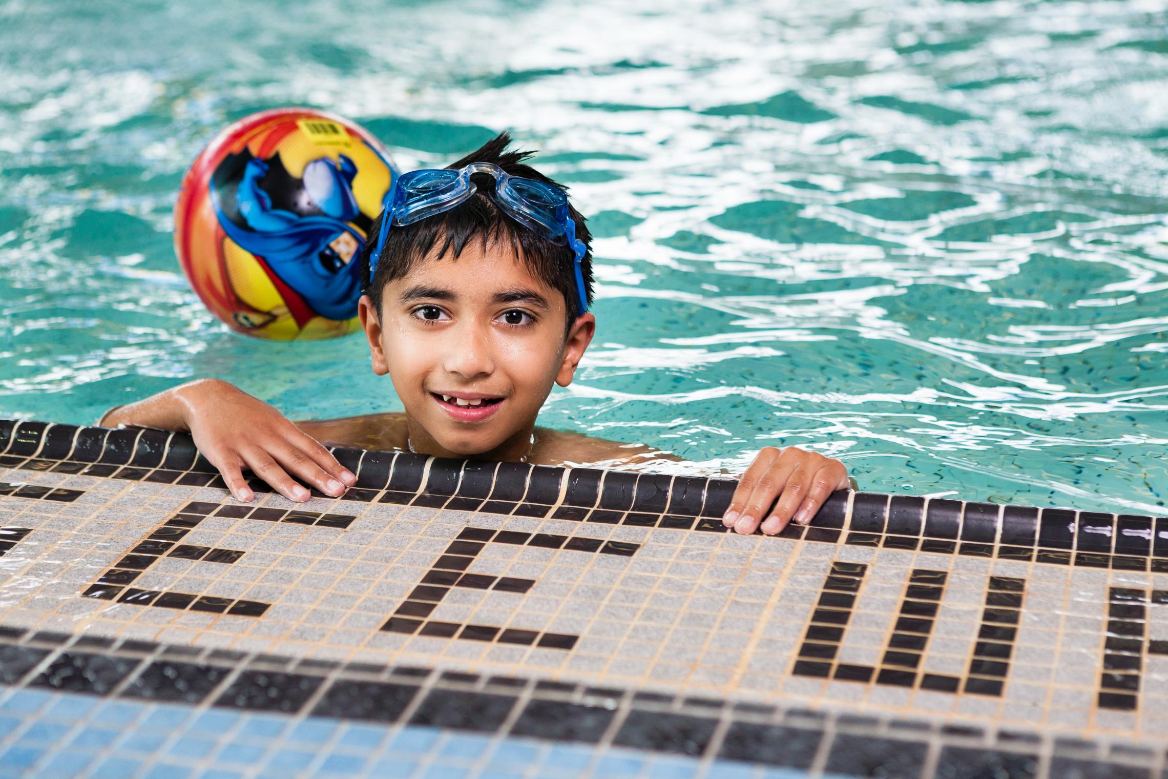 A young boy with goggles smiles and holds onto the side wall of a YMCA pool.