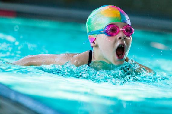A young girl in a multi-coloured swimming cap and goggles emerges from the water to take a breath while swimming.