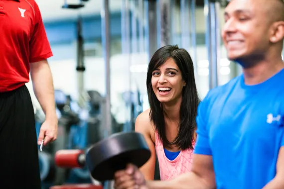 A woman in a pink tank top laughs with a man in a blue shirt lifting weights in a YMCA fitness facility.