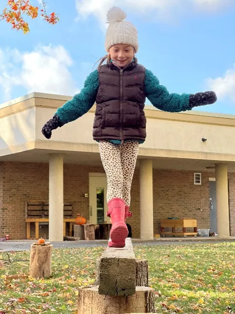 A girls wearing a white toque, black vest, and pink rain boots balances on a wooden log.