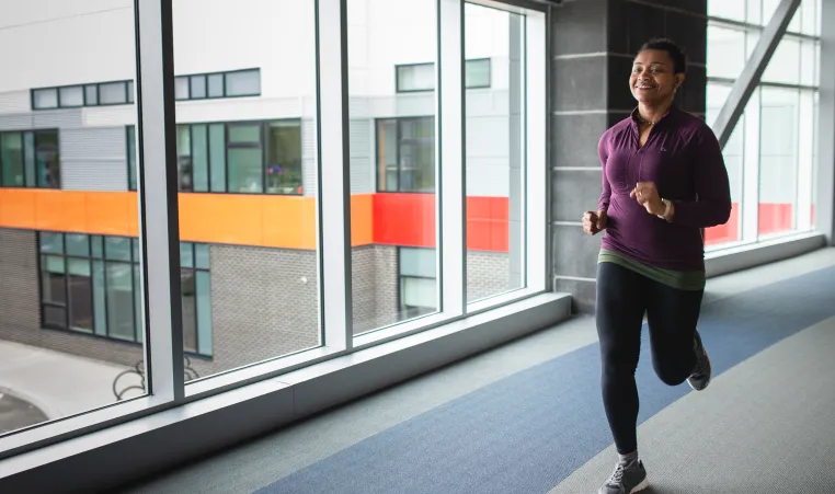 A woman in a purple shirt and black leggings jogs on an indoor walking track.