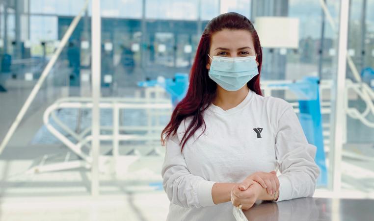 A young female staff member in a white shirt and mask smiles and poses at a health and fitness centre.