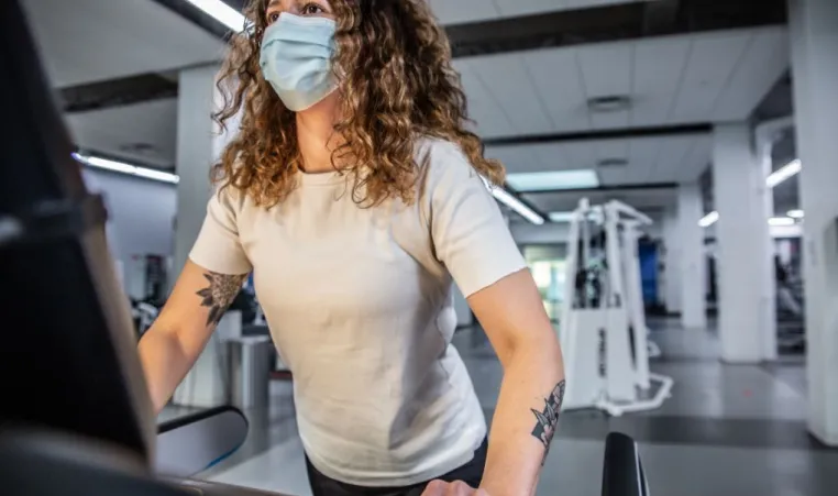 A woman with curly hair and tattoos on her arm, wearing a mask and white t-shirt, works out on a treadmill at the Y.