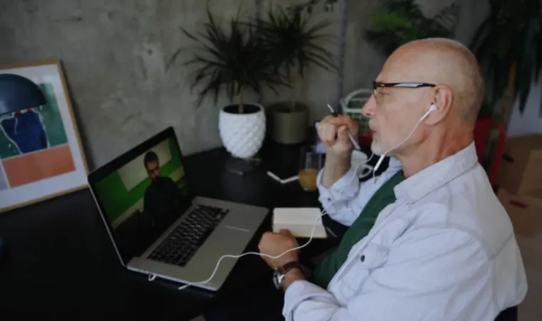 A man in a white button-up shirt with glasses and headphones sits at a computer desk while having a video call with another man on his laptop.