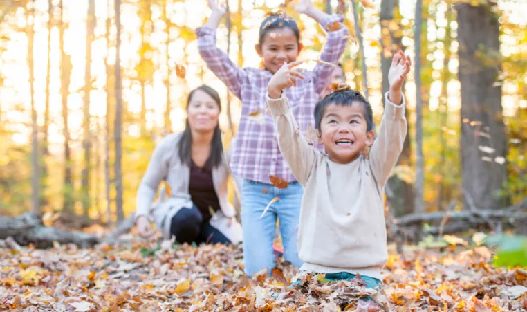 A young boy in a cream coloured sweater sits on the ground and smiles as he throws autumn leaves into the air. His sister and mom laugh and watch him from behind.