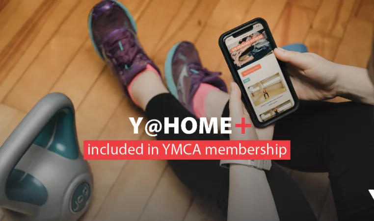 Y@HOME+ included in YMCA membership. Image: a woman in running shoes watches a workout on her mobile device next to a kettlebell.