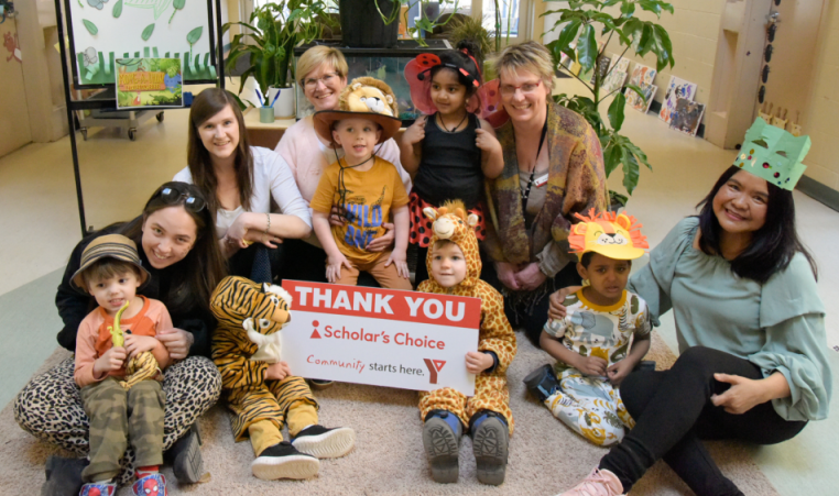 YMCA Staff pose with children from a Y Child Care Centre in costumes, holding a sign thanking Scholar's Choice for their sponsorship of Move-A-Thon.