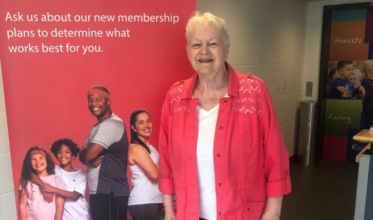 Barb smiles in front of a YMCA pop-up sign wearing a red shirt and black pants.