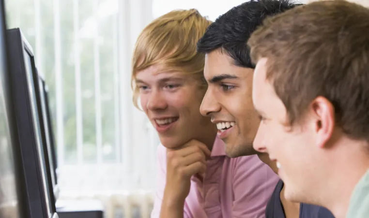 Three adolescents laugh as they play a game online in front of computer monitors.