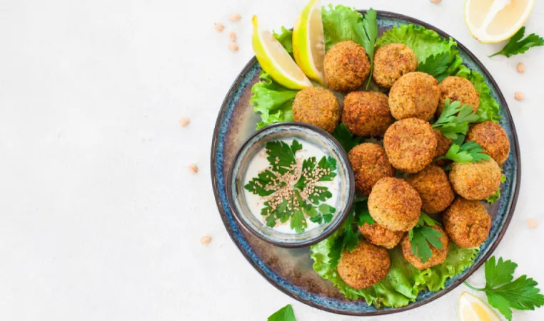 A blue bowl of falafels on a bed of lettuce with a side sauce.
