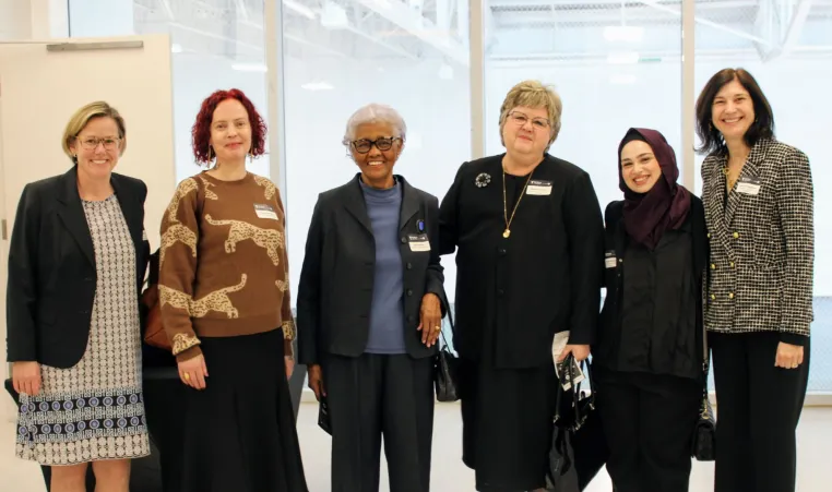 A group of six women standing together indoors, smiling at the camera. They are dressed in a mix of professional and semi-formal attire, wearing name tags. The background features large windows with a bright, open space behind them.