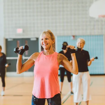An older woman in a pink tank top smiles and lifts weights as part of a group fitness class.