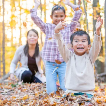 A young boy in a cream coloured sweater sits on the ground and smiles as he throws autumn leaves into the air. His sister and mom laugh and watch him from behind.