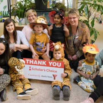 YMCA Staff pose with children from a Y Child Care Centre in costumes, holding a sign thanking Scholar's Choice for their sponsorship of Move-A-Thon.