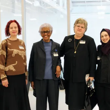 A group of six women standing together indoors, smiling at the camera. They are dressed in a mix of professional and semi-formal attire, wearing name tags. The background features large windows with a bright, open space behind them.