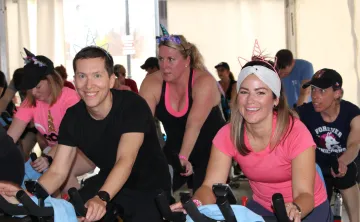 A group of people in pink and black shirts cycle on stationery bikes at a SWEAT for Strong Kids event.