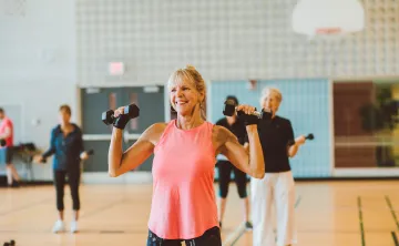 An older woman in a pink tank top smiles and lifts weights as part of a group fitness class.