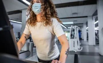 A woman with curly hair and tattoos on her arm, wearing a mask and white t-shirt, works out on a treadmill at the Y.