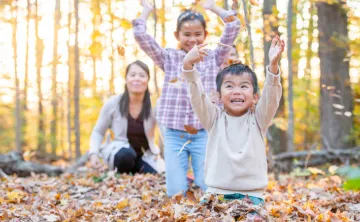 A young boy in a cream coloured sweater sits on the ground and smiles as he throws autumn leaves into the air. His sister and mom laugh and watch him from behind.