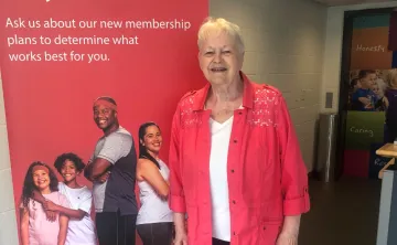 Barb smiles in front of a YMCA pop-up sign wearing a red shirt and black pants.