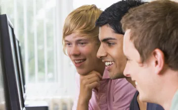 Three adolescents laugh as they play a game online in front of computer monitors.
