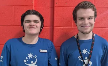 Two young men with light skin and short brown hair stand side by side in front of a red brick wall, smiling at the camera. They are both wearing matching blue t-shirts with a white logo featuring a stylized person, stars, and swirls. The man on the left has a name tag that reads 'Noah' and 'Project Search Team.' The man on the right wears a black lanyard with red 'Y' logos.