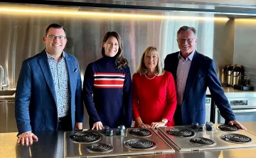 A group of four professionally dressed people stands behind a stainless steel kitchen counter with electric stovetops. They are smiling at the camera in a modern, well-lit kitchen with reflective metal surfaces, a large ventilation hood, and various kitchen appliances in the background.