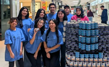 A group of nine young volunteers wearing matching blue t-shirts with "The Inn of The Good Shepherd" logo stand together in a shopping mall, smiling at the camera. They are positioned next to a large, neatly stacked display of canned food, primarily tuna and chickpeas. One volunteer in the front row holds up a can. In the background, shoppers walk through the mall, and a sign for the YMCA Canned Food Drive is visible.