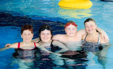 A family of four (two women, a girl and boy) smile and hug in a YMCA pool.